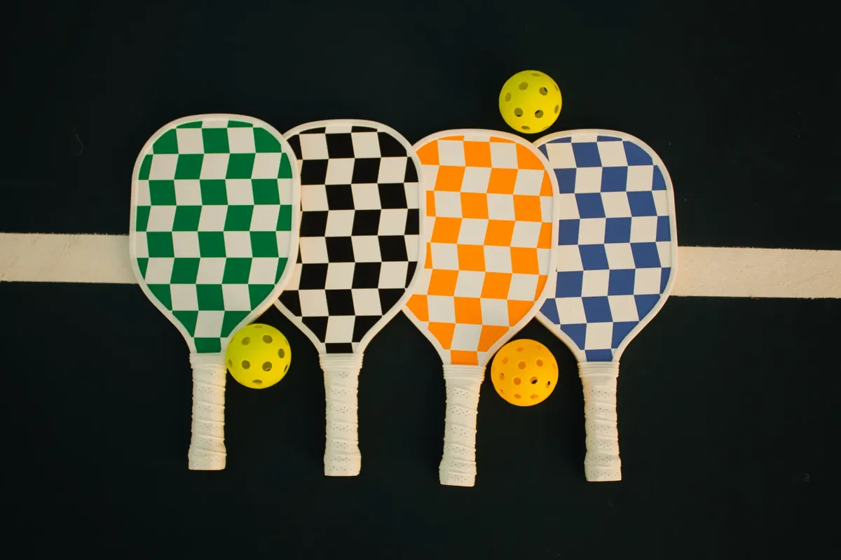 Four checkerboard-patterned pickleball paddles with wiffle balls on a sports court.