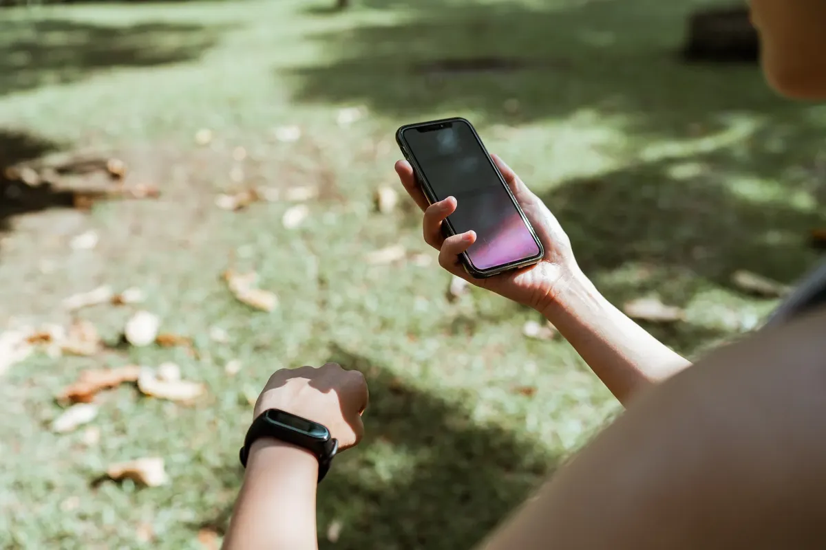 From above crop unrecognizable female wearing fitness tracker browsing modern mobile phone with blank screen while resting in green park