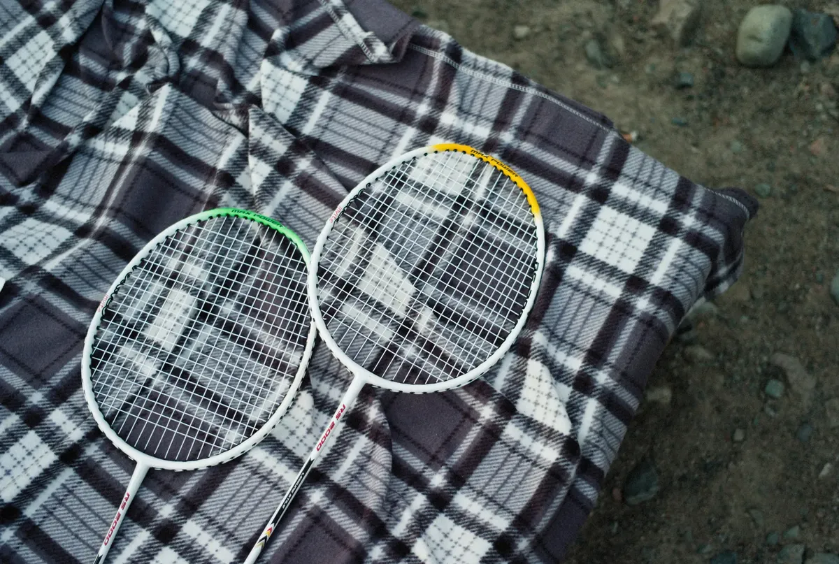 Two badminton rackets resting on a checkered picnic blanket outdoors, ideal for sports and leisure themes.