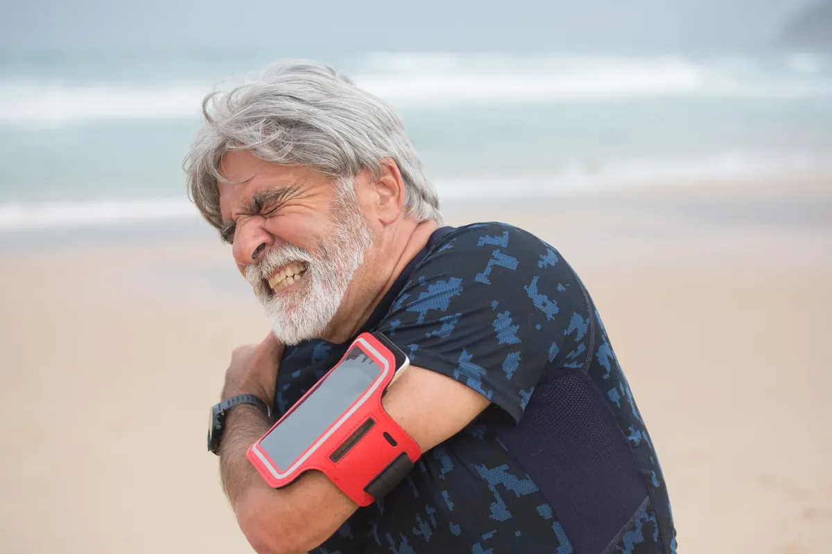 Elderly man grimacing from shoulder pain during seaside walk, showcasing health issue outdoors.